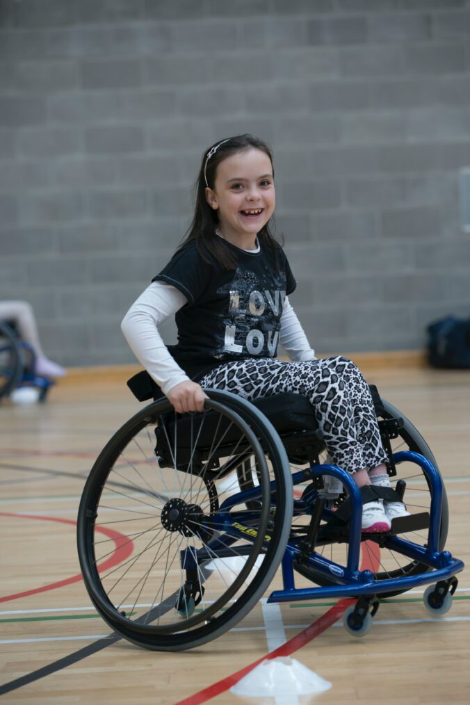 A young girl sits in a wheelchair in a gymnasium. She smiles at the camera.