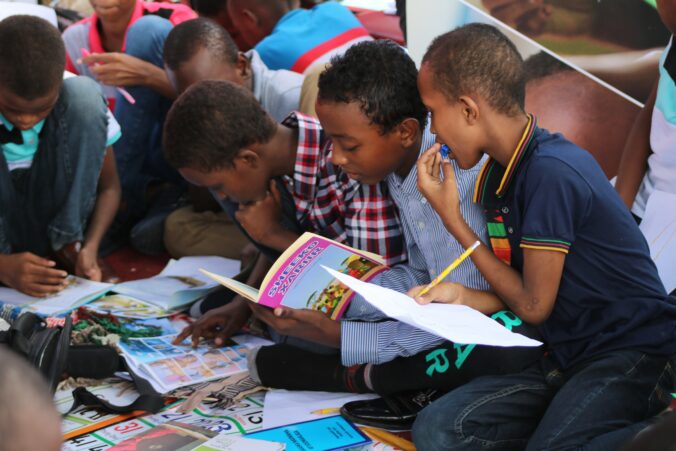 Children sit on the group looking at books and magazines. The child closest to the camera has a pencil and paper in his hand.